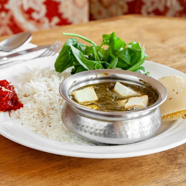 Indian dish with palak paneer in a metal bowl, rice, chutney, and papadum on a white plate.
