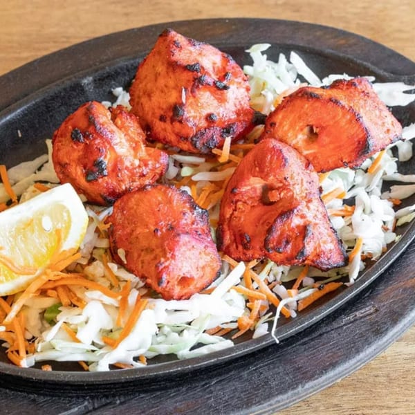 Five pieces of tandoori chicken tikka on a bed of shredded cabbage and carrots with a lemon wedge.