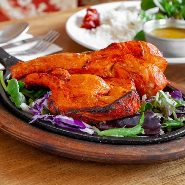 Two pieces of tandoori chicken on a sizzling platter with a side of salad, rice, and sauce.